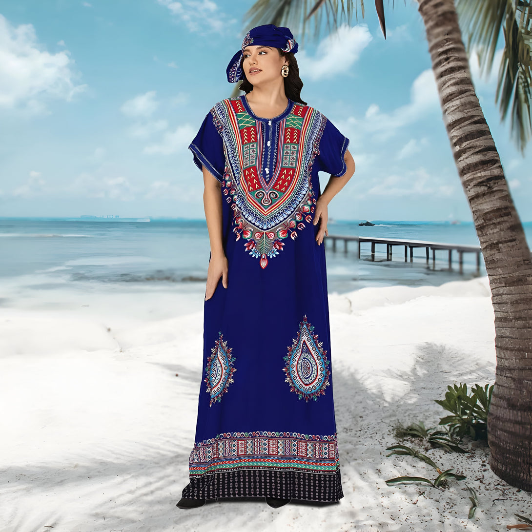 Une femme portant une robe longue bleue avec des motifs ethniques multicolores, un foulard assorti et des chaussures noires, debout sur la plage.