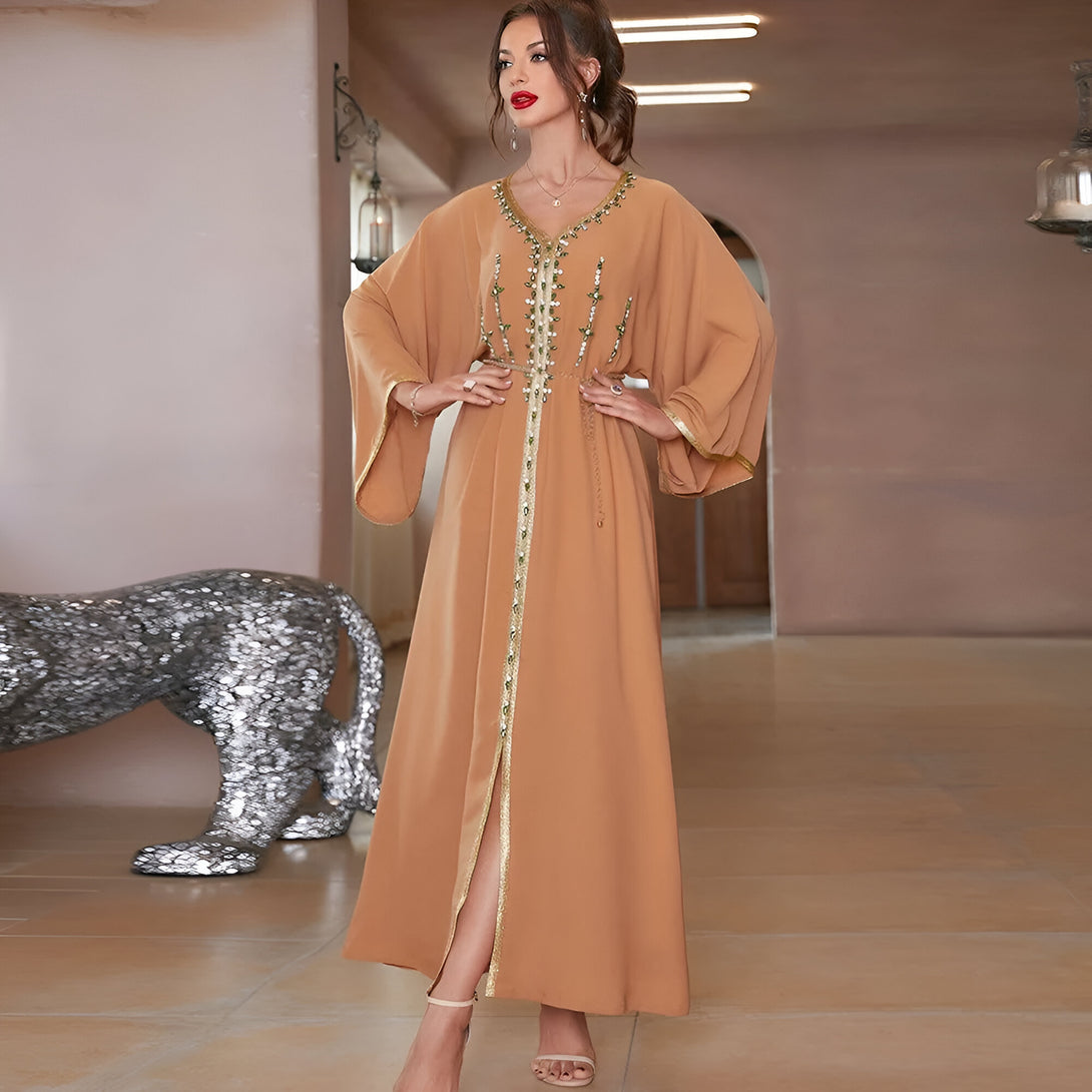 Une femme porte une longue robe beige à manches longues avec une ceinture et des détails brodés.