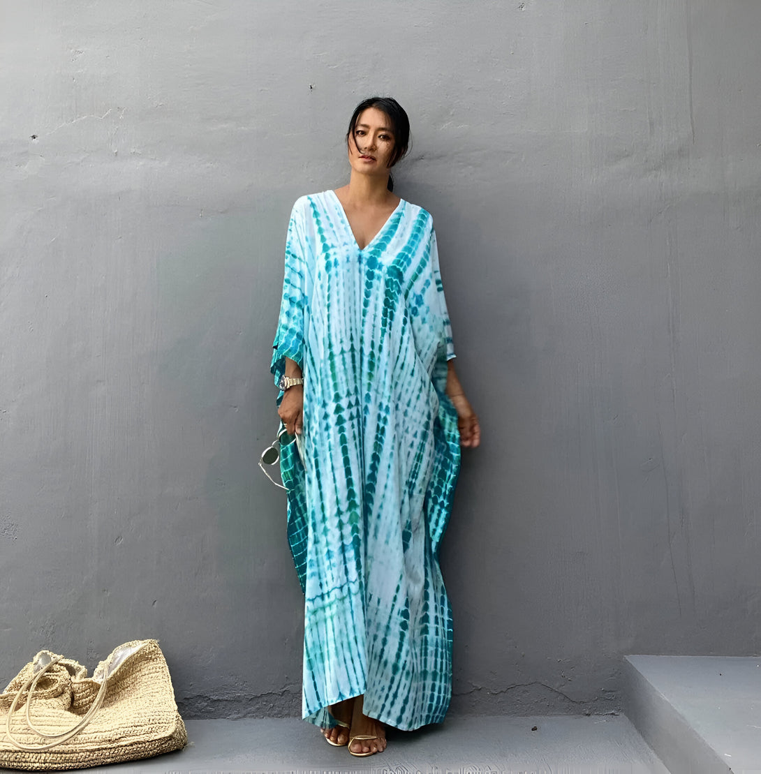 Une femme portant une longue robe bleu turquoise à imprimé tie-dye. La robe est ample et a des manches 3/4. La femme a les cheveux noirs et porte des sandales dorées. Elle se tient debout devant un mur gris.