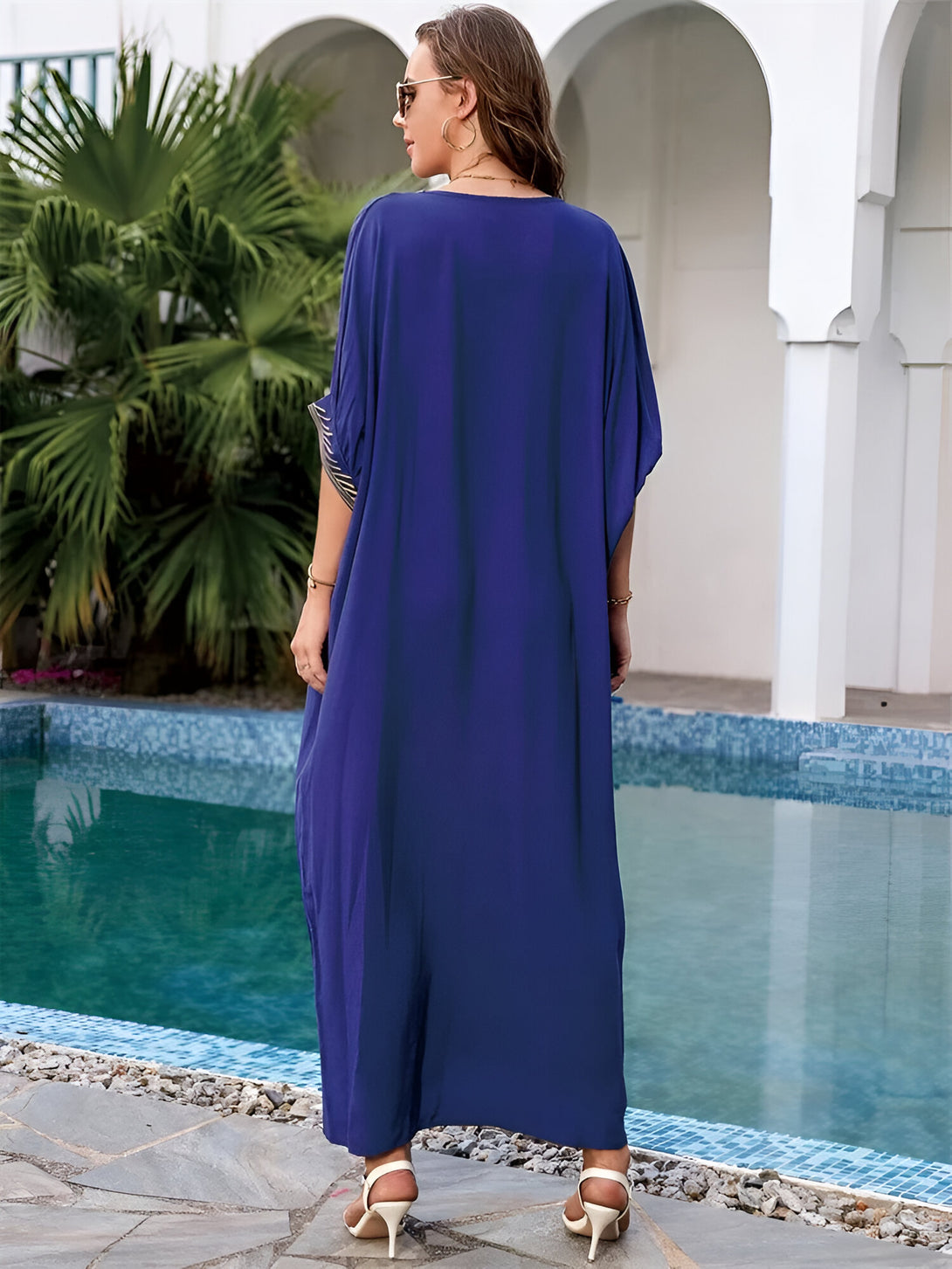 Une femme portant une longue robe bleue avec des détails dorés, debout près d'une piscine. La robe est ample et fluide, idéale pour les journées chaudes.