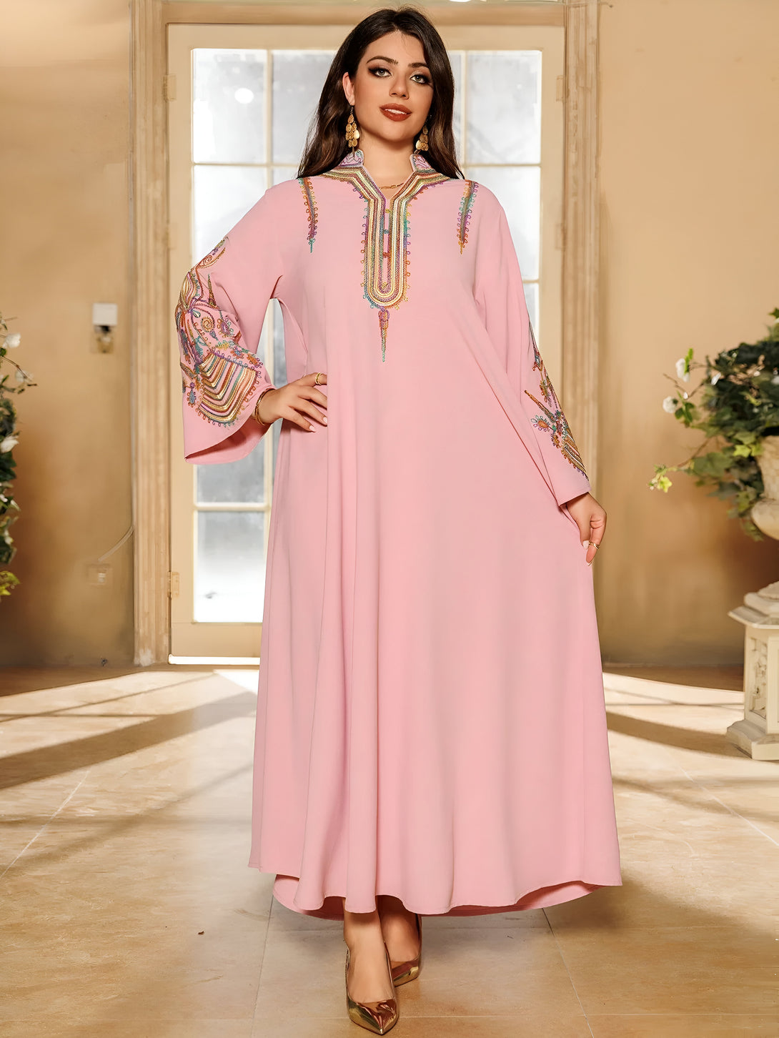 Une femme porte une robe rose longue avec des manches longues brodées de motifs multicolores. La robe est ample et fluide.