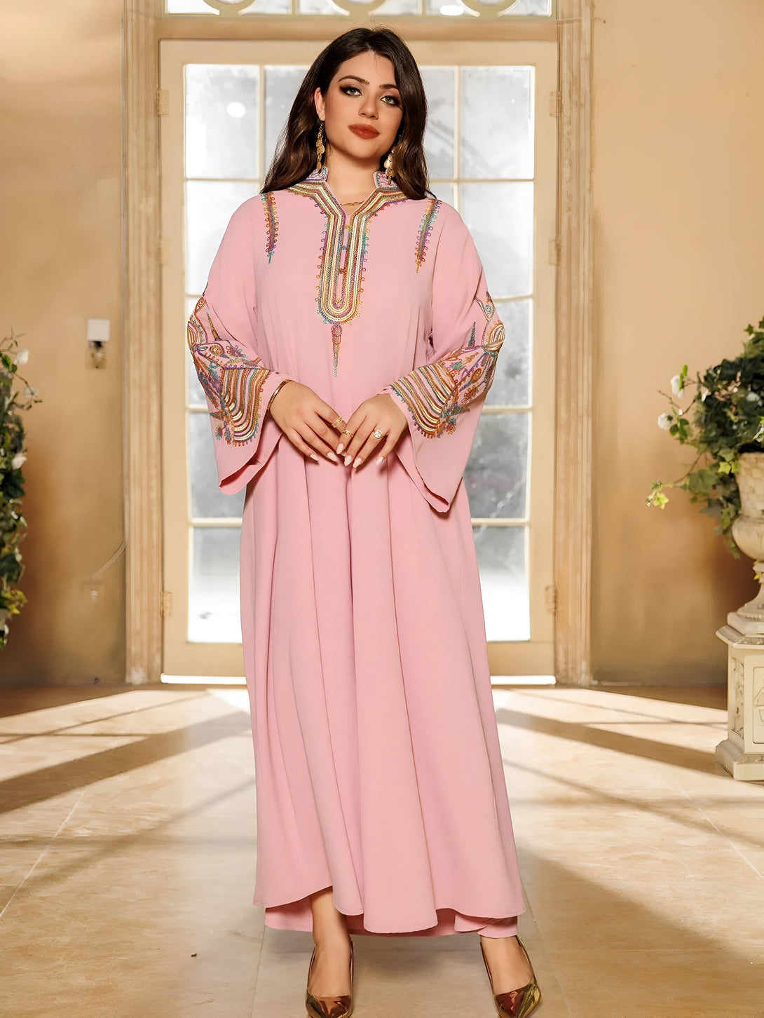 Une femme porte une robe rose à manches longues brodée de motifs multicolores. La robe est ample et tombe jusqu'aux pieds.