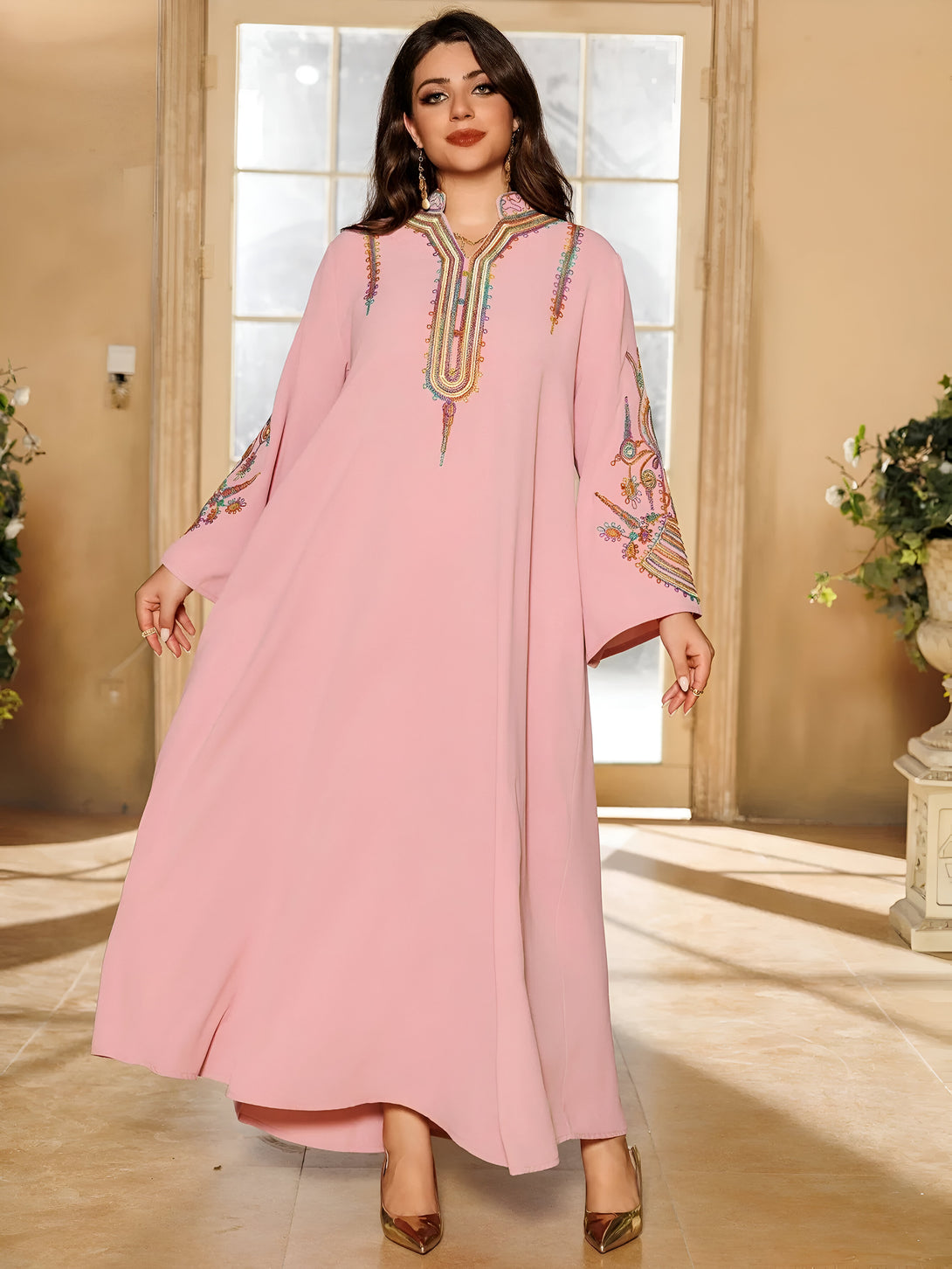 Une femme porte une longue robe rose avec des broderies colorées. La robe a des manches longues et un col haut. Elle porte des chaussures dorées.