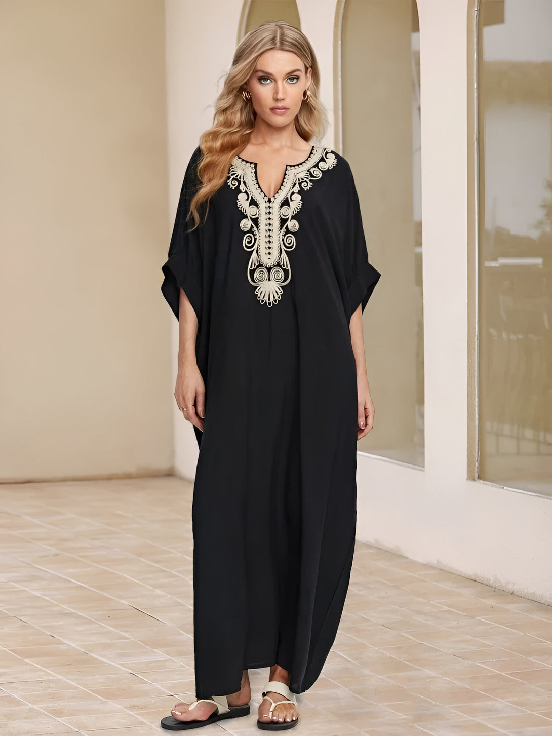 Une femme porte une longue robe noire à manches courtes avec des broderies dorées. La robe est ample et fluide, idéale pour les journées chaudes.