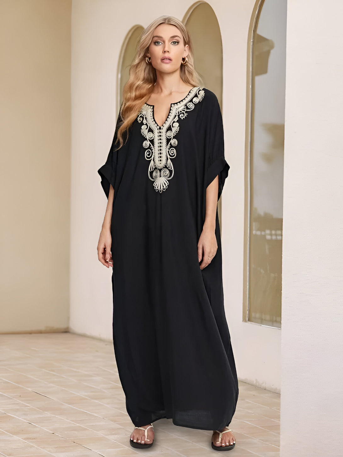 Une femme portant une robe noire longue avec des broderies dorées. La robe a un col en V et des manches courtes. La femme porte des sandales plates et a les cheveux blonds. La robe est parfaite pour les occasions spéciales ou pour un look élégant au quotidien.