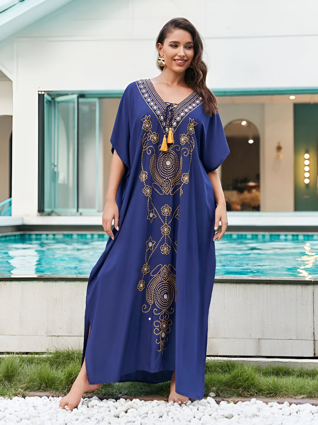 Une femme souriante porte une longue robe bleue à manches courtes ornée de motifs dorés. La robe est cintrée à la taille et comporte une encolure en V. La femme est debout devant une piscine.