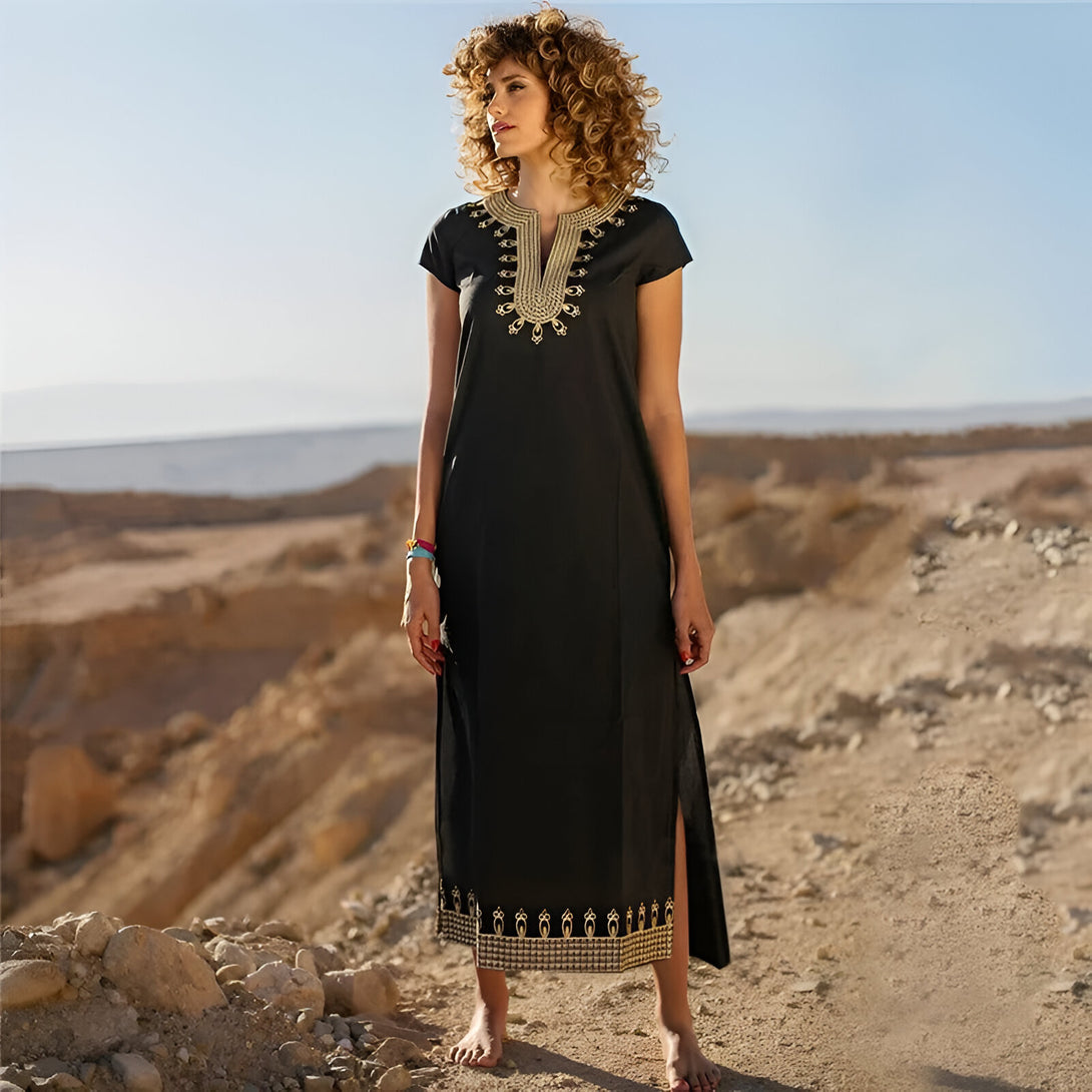 Une femme portant une robe noire ample avec des broderies dorées, dans le désert. Elle porte un vêtement musulman.