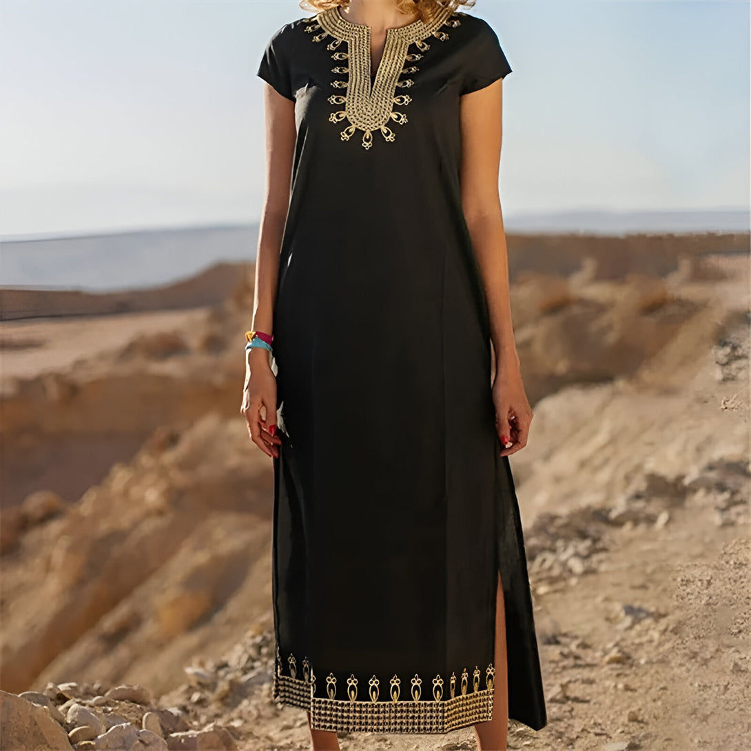 Une femme porte une robe noire longue avec des broderies dorées. La robe a des manches courtes et une encolure en V. Elle est debout dans un désert.