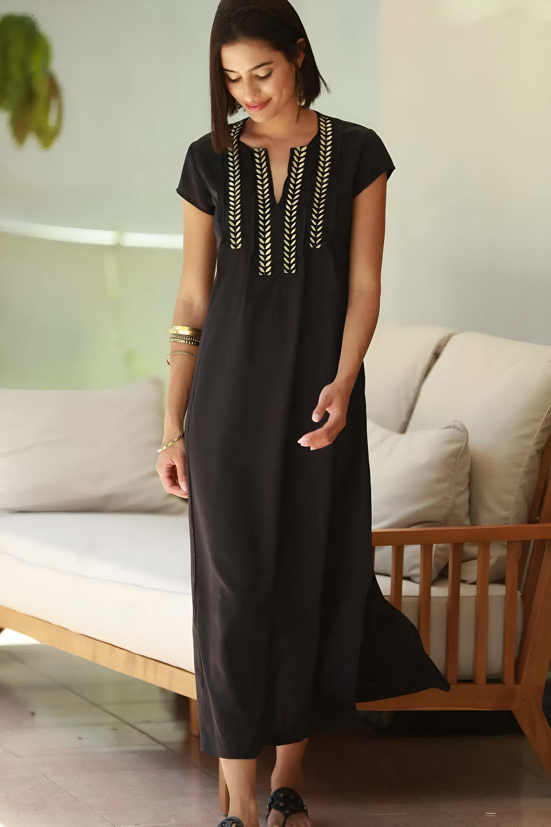 Une femme portant une robe noire à manches courtes avec des broderies dorées. La robe a une encolure en V et une coupe ample. La femme est debout devant un canapé en bois.