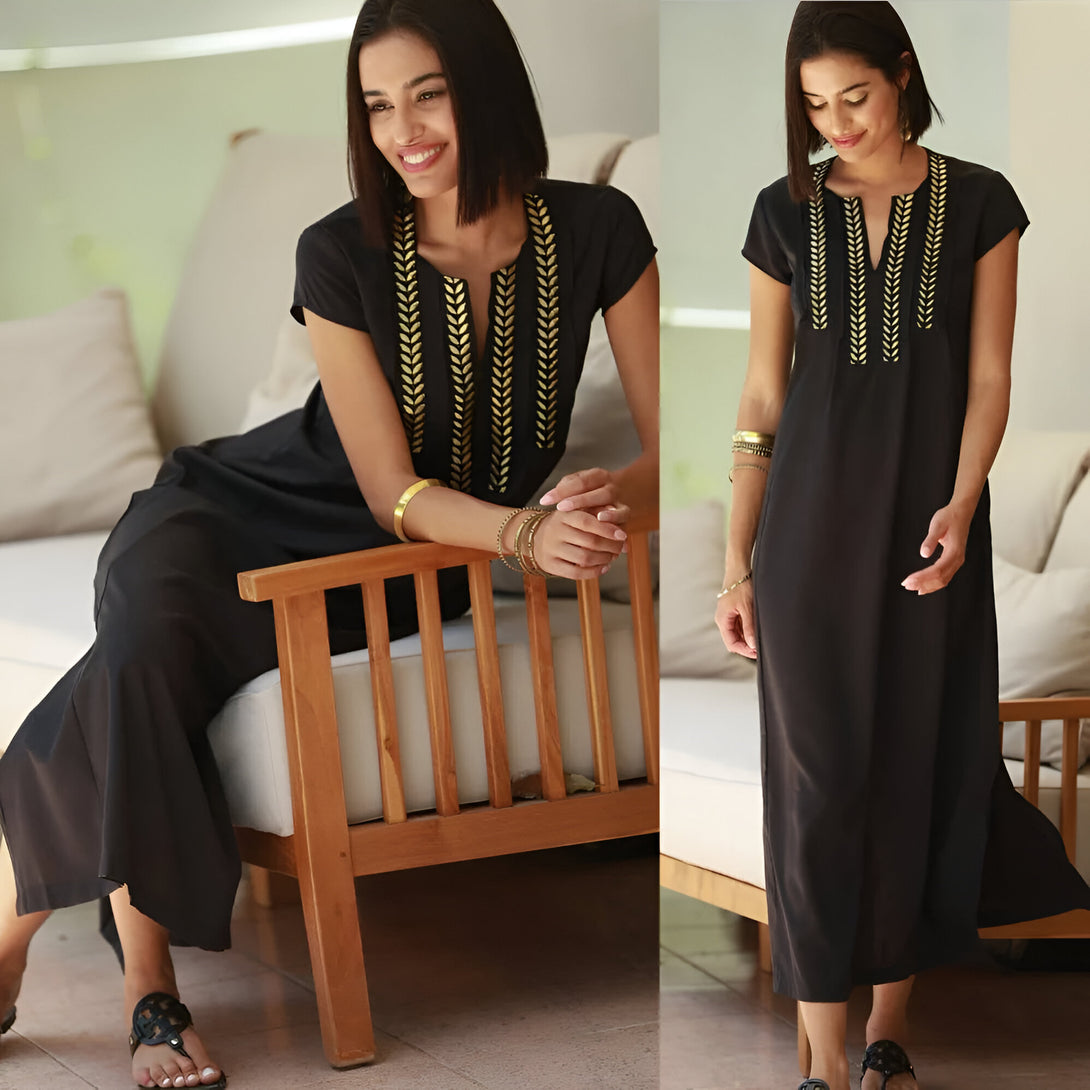 Une femme porte une robe noire longue avec des détails dorés. La robe a un col en V et des manches courtes. La femme est assise sur un canapé en bois et sourit à la caméra. Elle porte également des bracelets dorés.  Robe musulmane élégante et confortable.