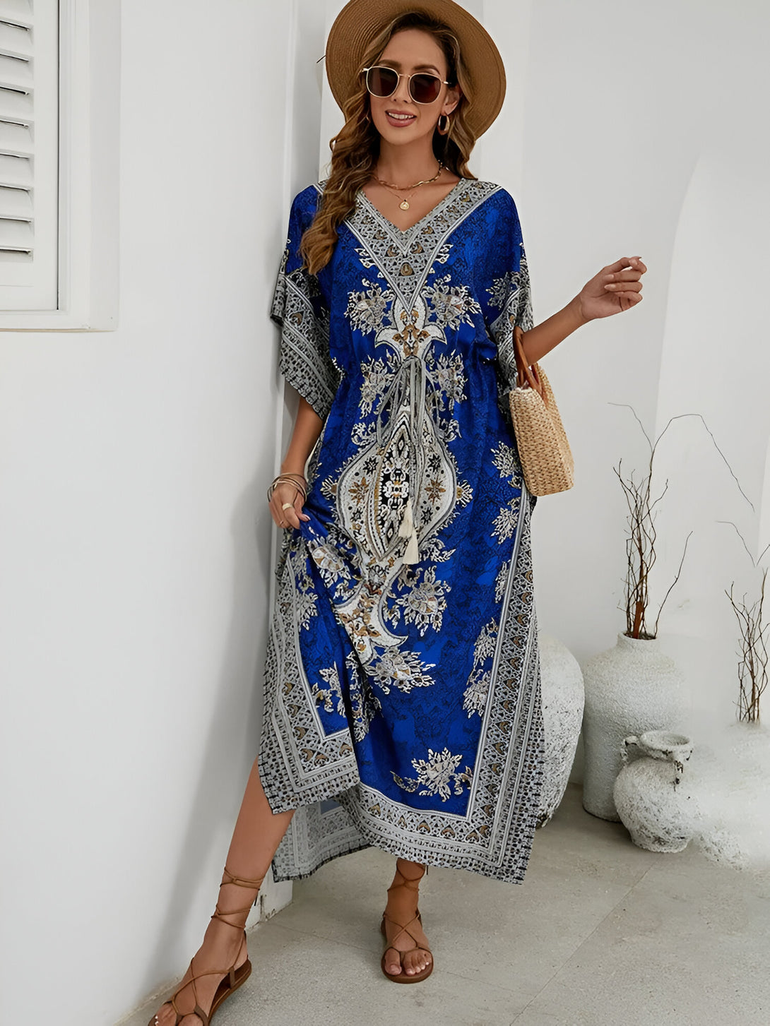 Une femme porte une longue robe bleue à manches courtes avec un motif floral. La robe a une ceinture à la taille et une fente sur le côté. Elle porte également un chapeau de paille et des lunettes de soleil.