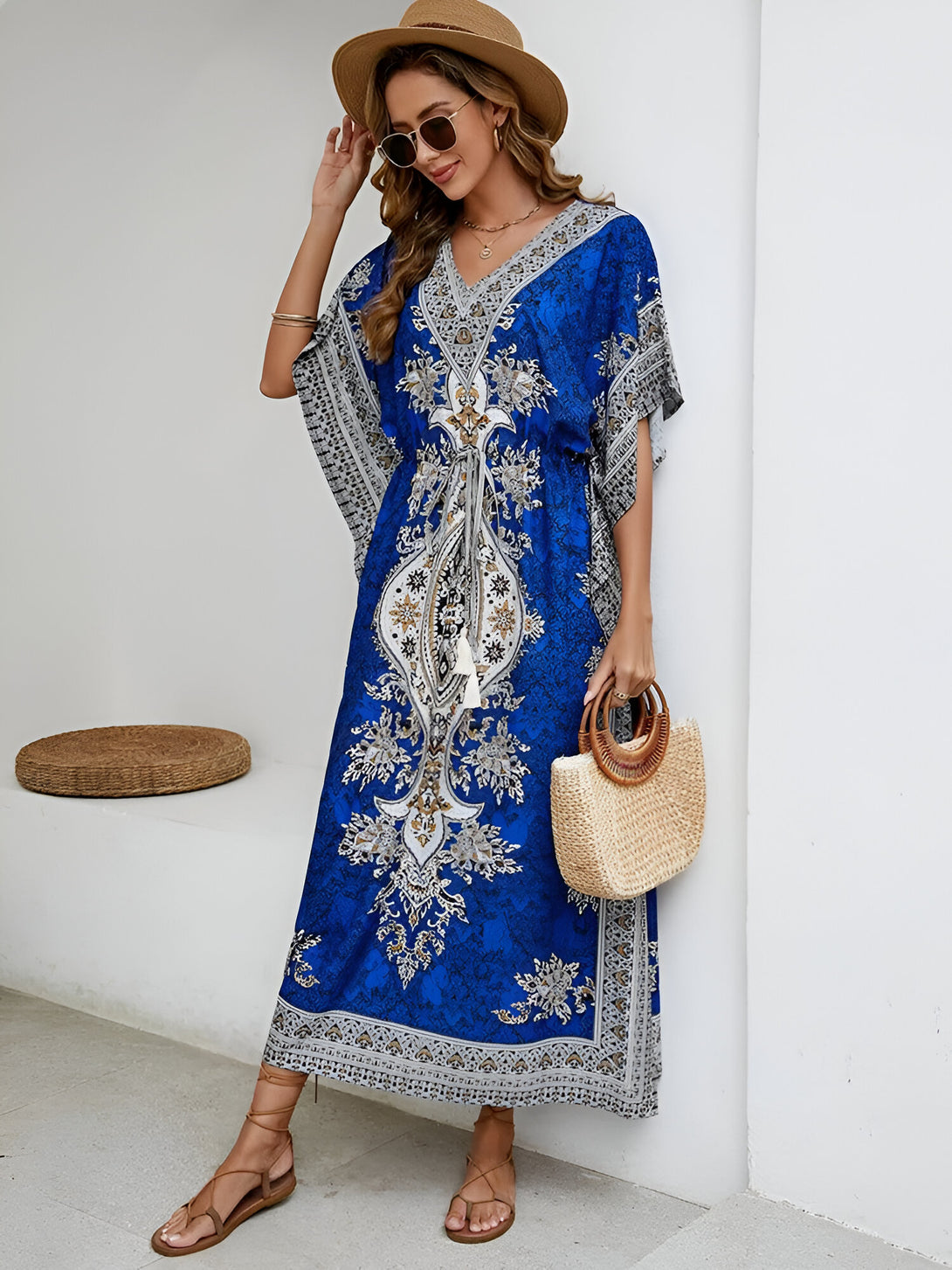 Une femme porte une longue robe bleue à motifs floraux. Elle porte un chapeau de paille et des lunettes de soleil. Elle tient un sac à main en osier.  La robe est parfaite pour l'été et s'adapte à de nombreuses occasions.