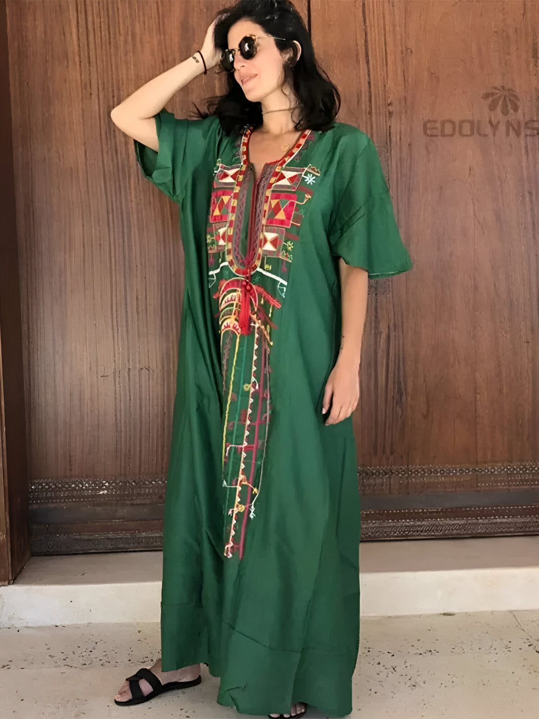 Une femme portant une longue robe verte avec des broderies colorées. La robe est ample et a des manches courtes. La femme porte des lunettes de soleil et a les cheveux noirs.