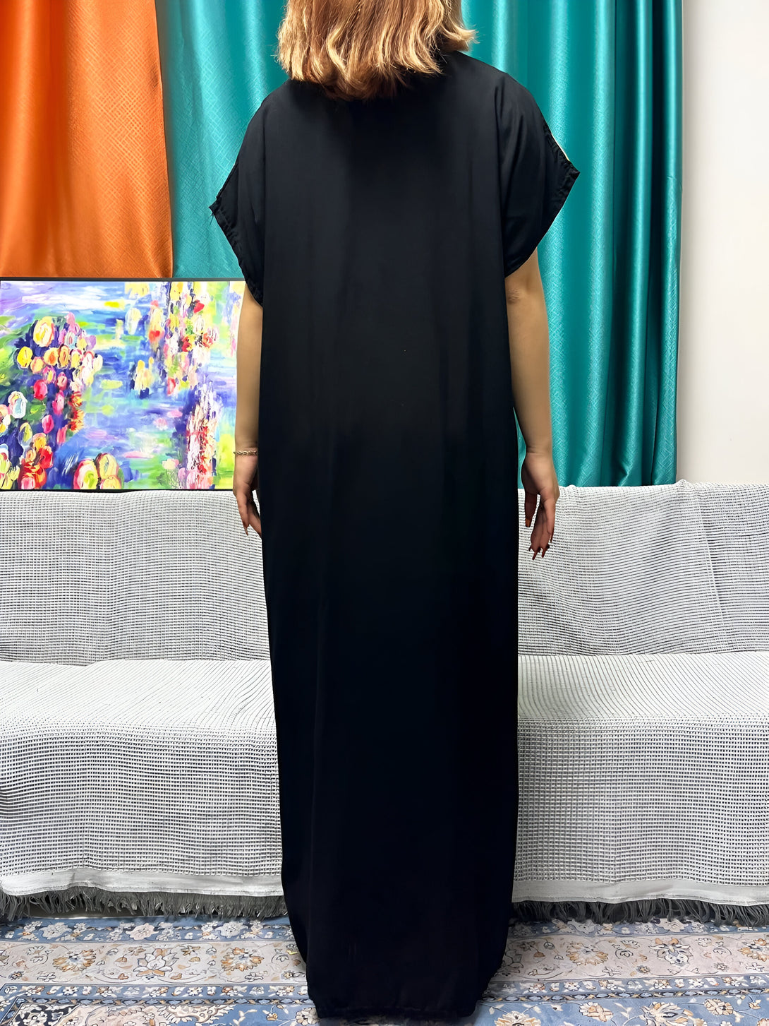 Une femme portant une longue robe noire ample avec des manches courtes. La robe est de style musulman, avec un col rond et un tissu fluide. La femme est debout dans une pièce avec un canapé et un tapis.