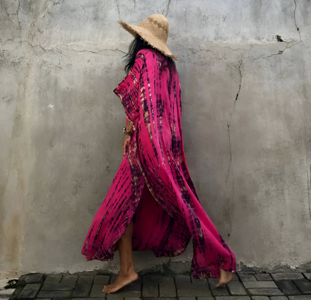 Une femme portant une longue robe rose à imprimé tie and dye, avec un chapeau de paille, se tient devant un mur en béton.
