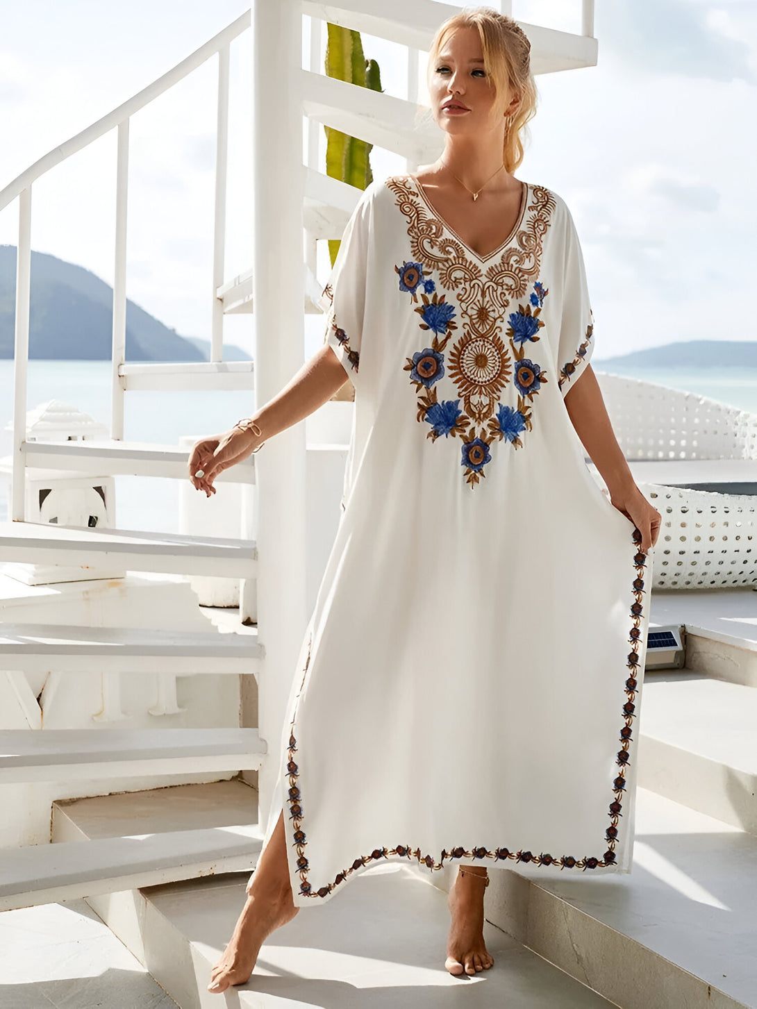 Une femme porte une longue robe blanche avec des broderies florales. La robe a un décolleté en V et des manches courtes. La robe est portée sur un fond de ciel bleu et d'eau bleue.