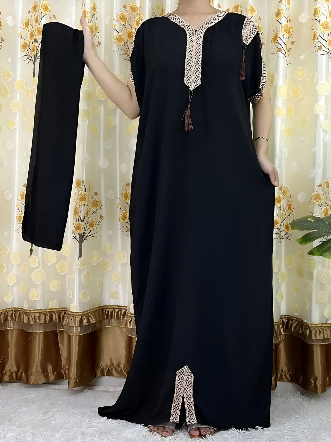 Une robe noire longue avec des manches courtes, ornée de dentelle beige et de franges, accompagnée d'un hijab noir.