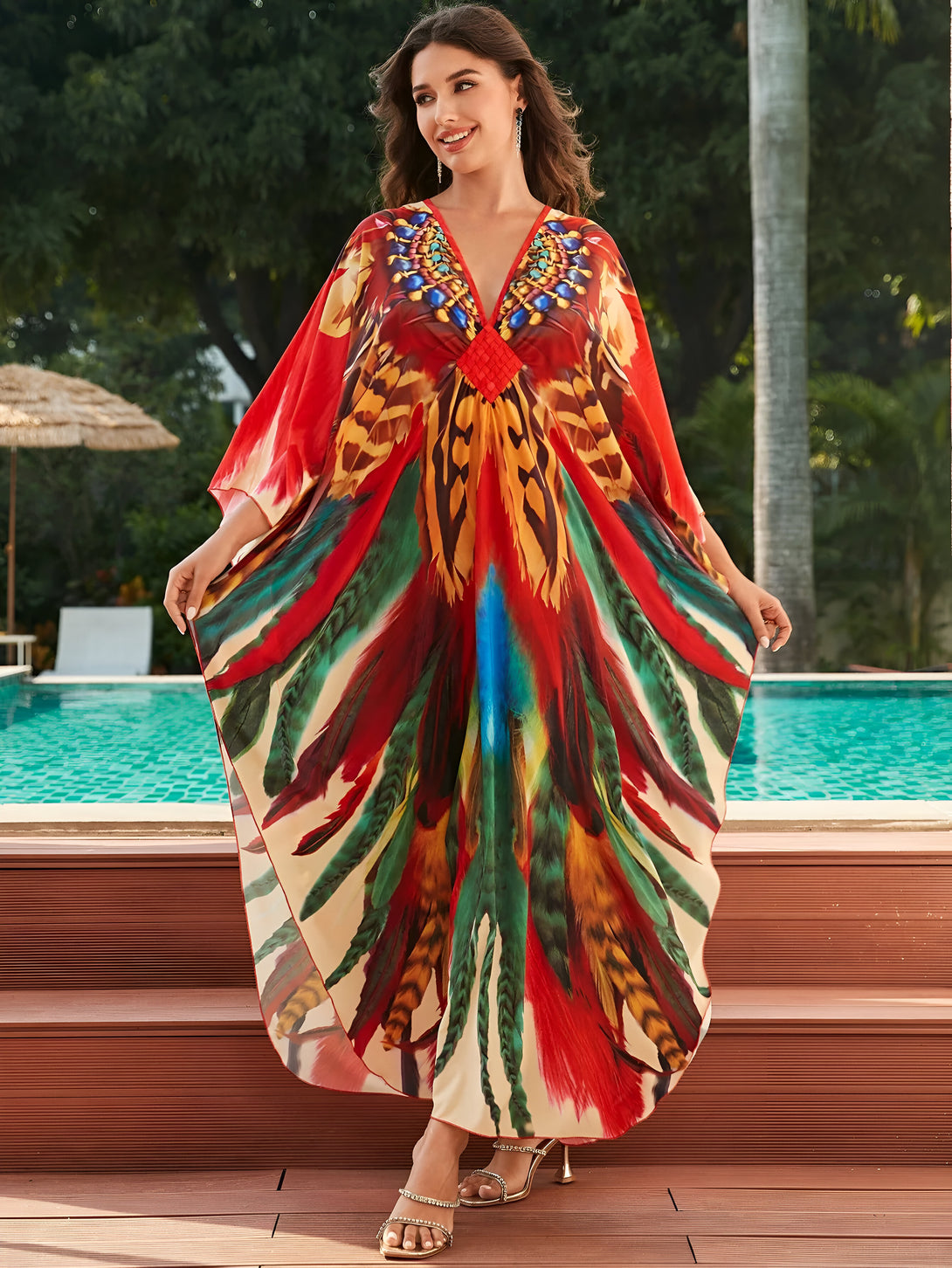 Une femme souriante porte une longue robe rouge à imprimé plumes colorées, debout près d'une piscine. La robe est parfaite pour les occasions spéciales ou pour se détendre à la plage.