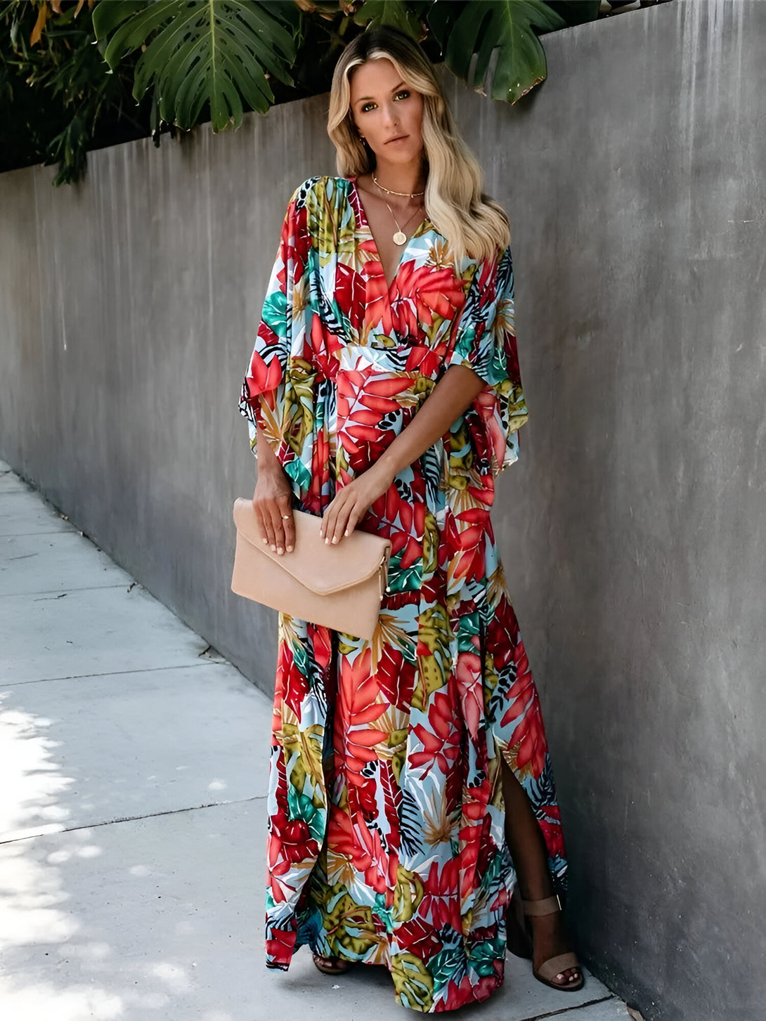 Une femme porte une longue robe à imprimé floral rouge, verte et bleue. La robe a des manches 3/4 et une fente sur le côté. La robe est parfaite pour l'été ou les occasions spéciales.