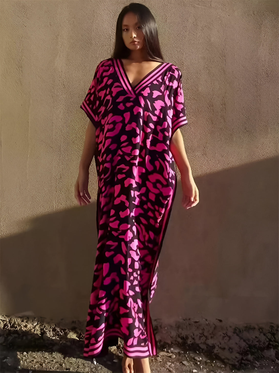 Une femme portant une longue robe rose à imprimé léopard avec un col en V. La robe a des manches courtes et est légèrement ample. La femme est debout devant un mur en béton.