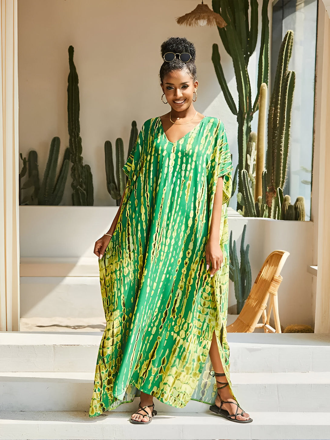 Une femme souriante porte une longue robe verte à imprimé tie-dye avec des manches courtes et des fentes sur les côtés. Elle porte des sandales et des lunettes de soleil. La robe est parfaite pour les occasions spéciales ou pour un look décontracté chic.