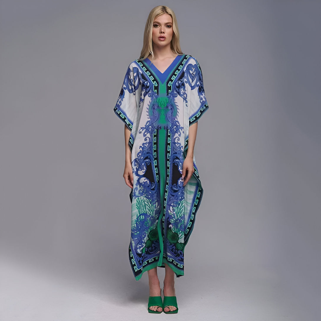 Une femme portant une longue robe blanche à manches courtes avec un motif bleu et vert. La robe a un col en V et une coupe ample. La femme porte des sandales vertes.