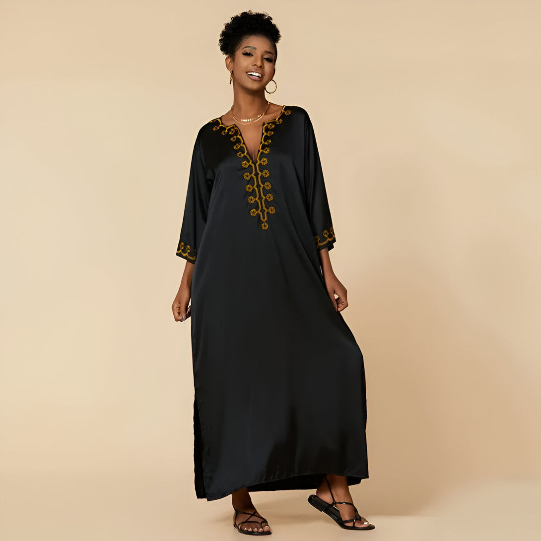 Une femme porte une robe noire longue avec des broderies dorées. La robe a une encolure en V et des manches trois-quarts. La femme porte des sandales noires.
