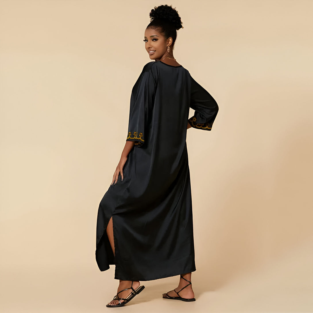 Une femme porte une robe longue noire élégante avec des détails dorés. La robe a des manches 3/4 et une fente sur le côté. Elle est parfaite pour les occasions spéciales ou les vêtements décontractés.