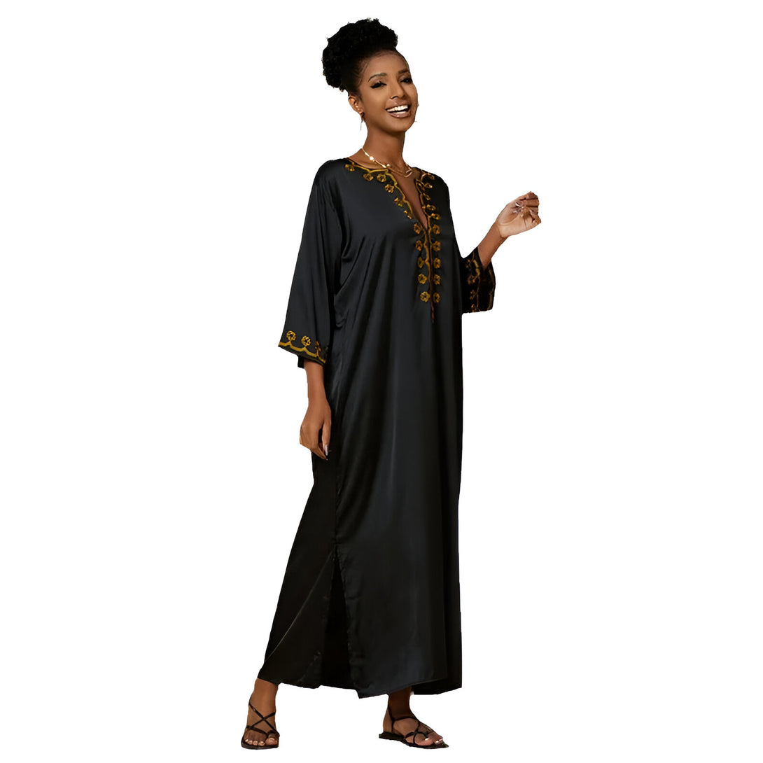 Une femme souriante portant une robe longue noire avec des broderies dorées. La robe a des manches longues et une fente sur le côté.