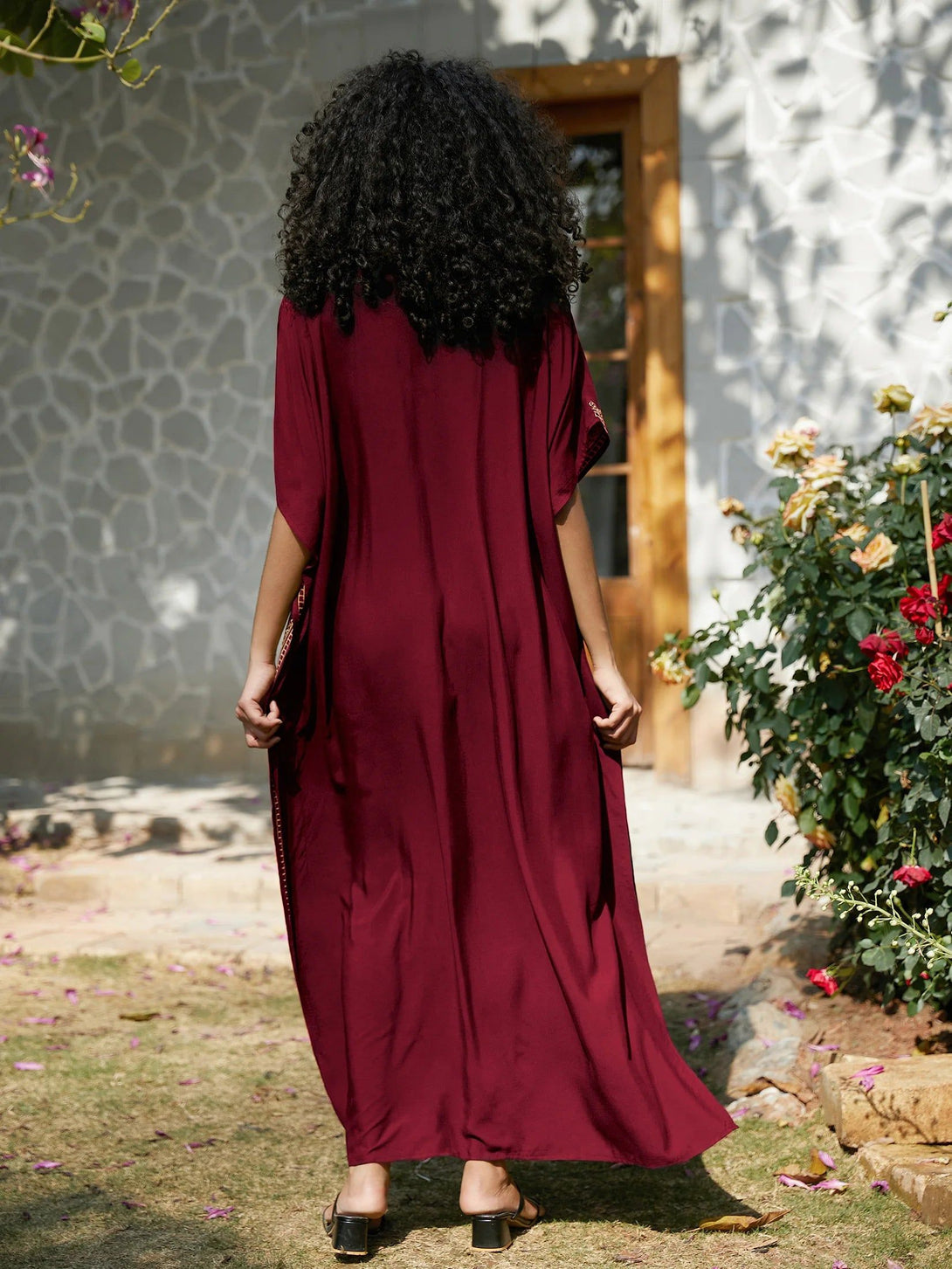 Une femme portant une longue robe rouge à manches courtes, debout dans un jardin. La robe est fluide et ample, avec un col rond. La femme est de dos et ses cheveux sont longs et bouclés. Elle porte des sandales noires.