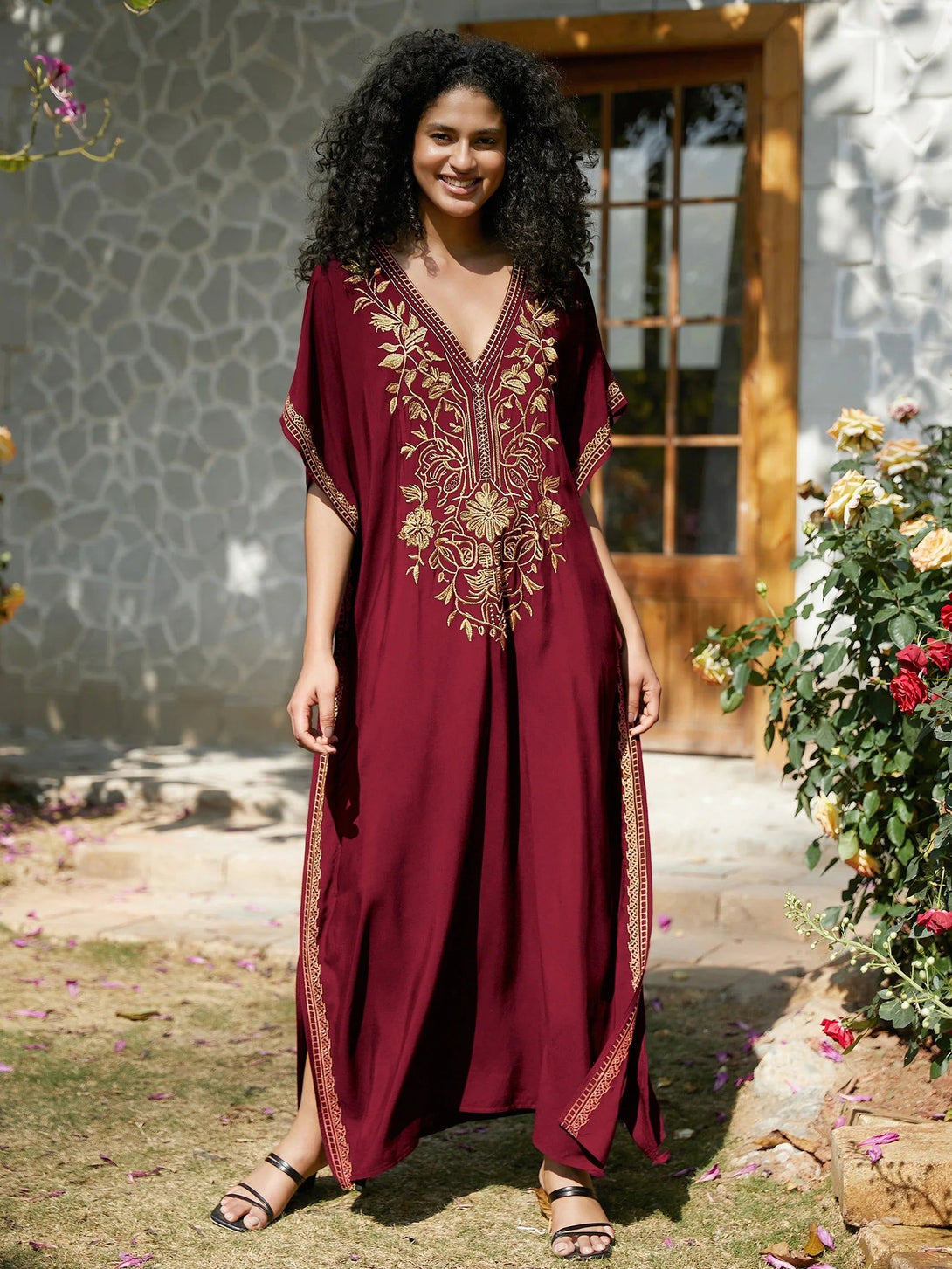 Une femme souriante portant une longue robe rouge avec des broderies dorées, debout dans un jardin. La robe est idéale pour les occasions spéciales et est disponible dans notre collection de vêtements musulmans.