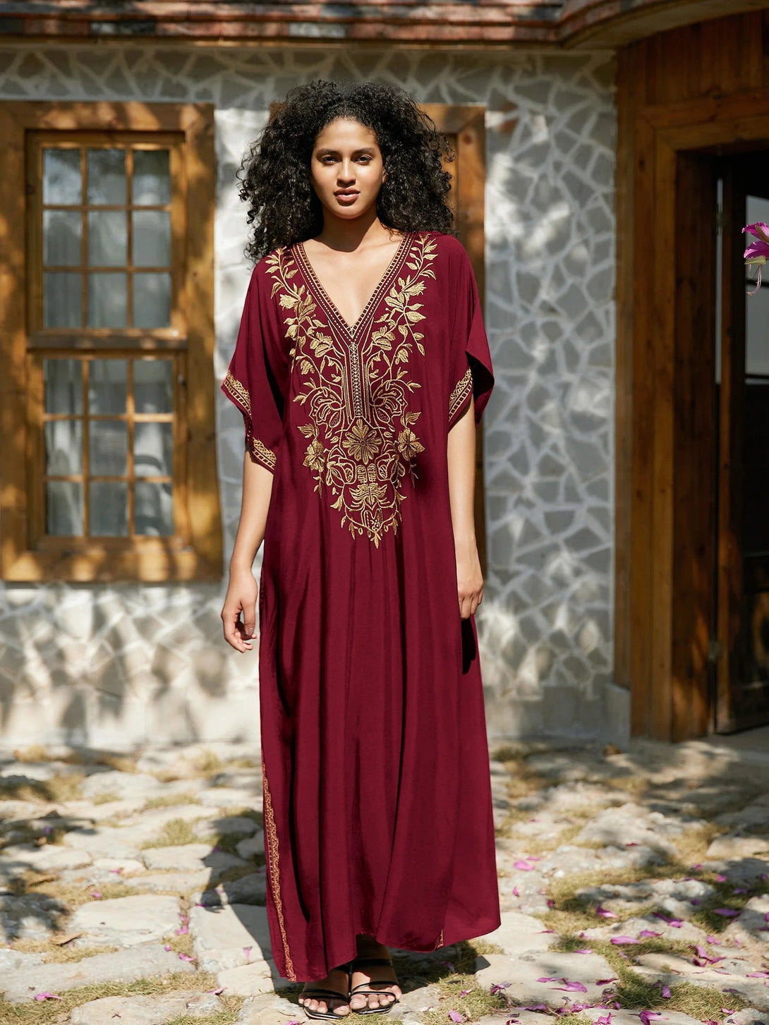 Une femme portant une robe longue bordeaux avec des broderies dorées. La robe a un col en V et des manches courtes. La femme est debout devant un bâtiment en pierre avec un jardin.