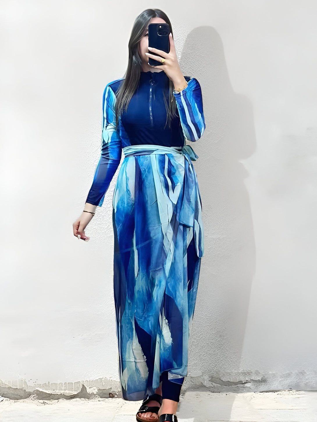 Une femme porte une robe de bain musulmane bleue à motifs abstraits. La robe a une fermeture éclair sur le devant et une ceinture à nouer. La femme porte des sandales et tient un téléphone dans sa main.