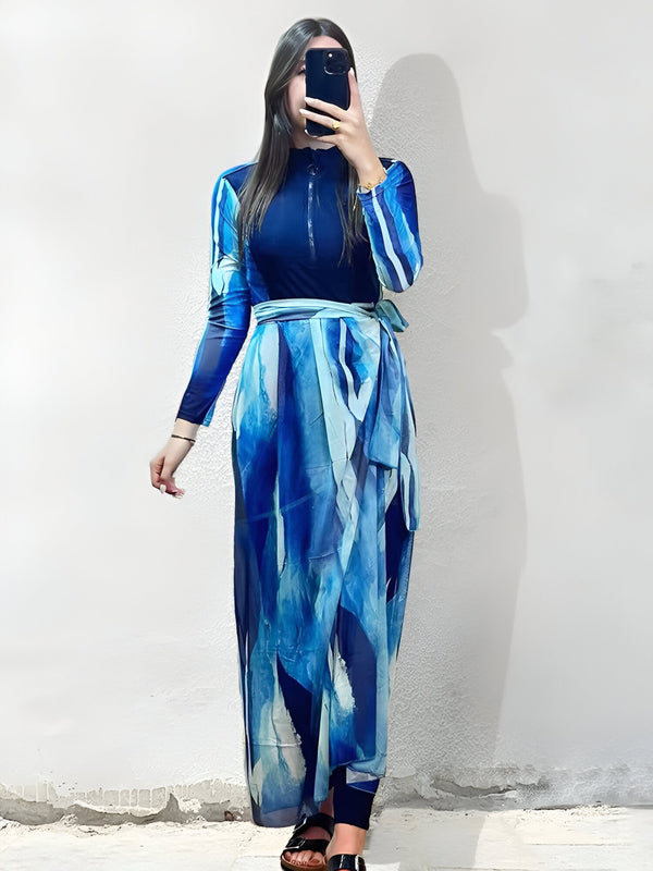 Une femme porte une robe de bain musulmane bleue à motifs abstraits. La robe a une fermeture éclair sur le devant et une ceinture à nouer. La femme porte des sandales et tient un téléphone dans sa main.