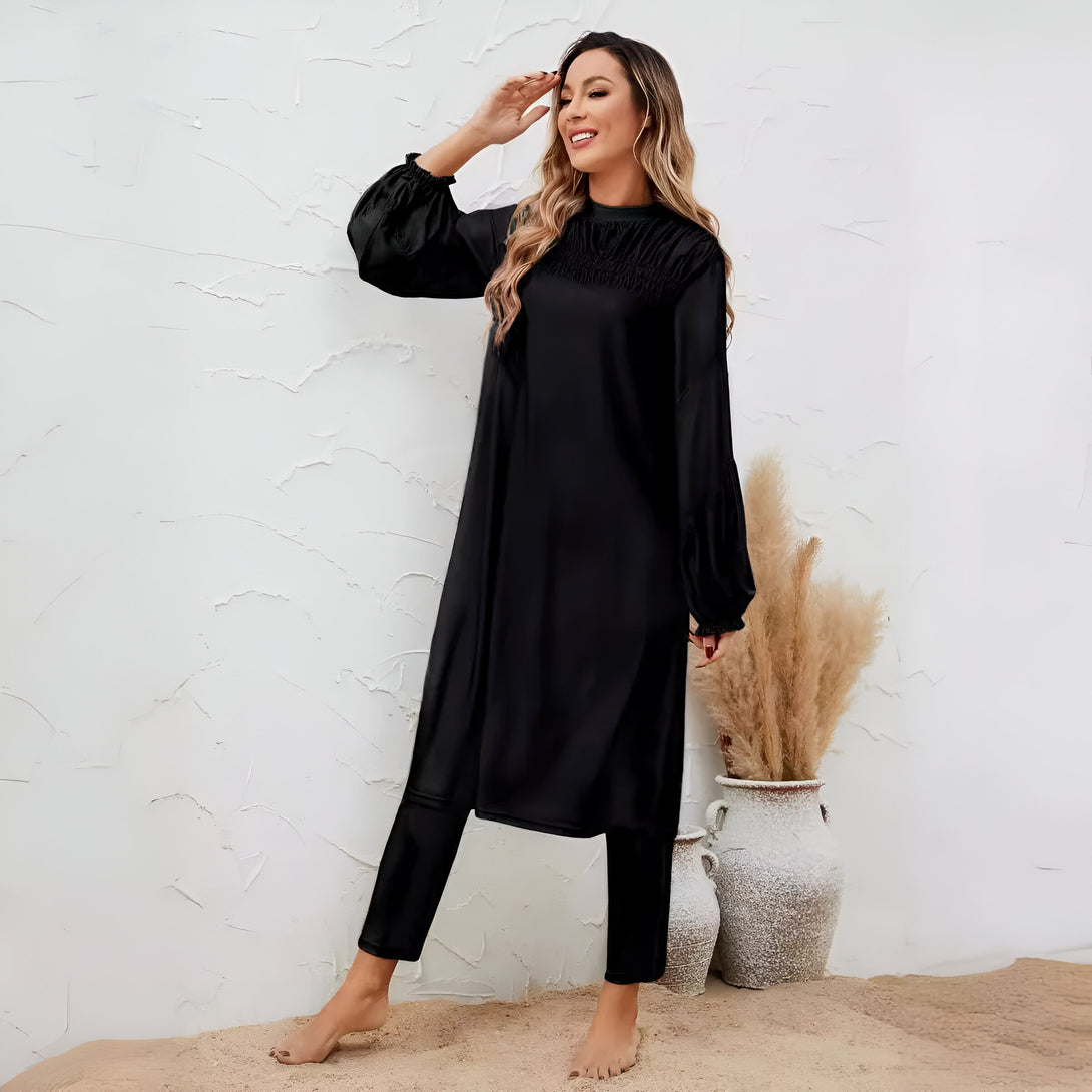 Une femme porte une robe noire ample avec des manches bouffantes et un pantalon noir assorti. Elle se tient debout sur un fond blanc, souriant à la caméra.