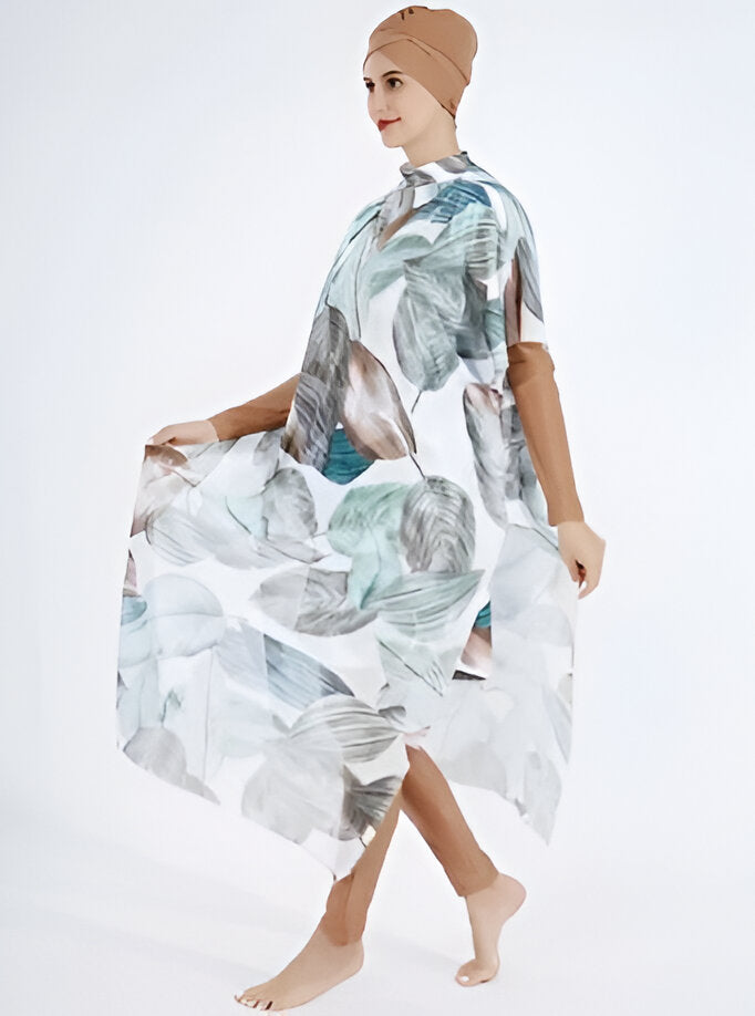 Une femme portant une robe longue de plage musulmane avec un imprimé de feuilles. La robe est blanche avec des feuilles vertes et grises. La femme porte un foulard marron sur la tête.