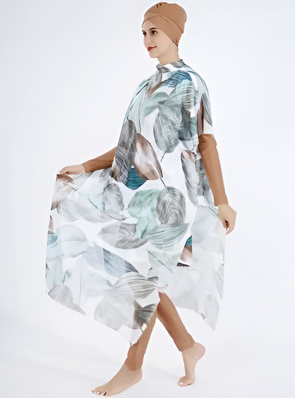 Une femme portant une robe longue de plage musulmane avec un imprimé de feuilles. La robe est blanche avec des feuilles vertes et grises. La femme porte un foulard marron sur la tête.