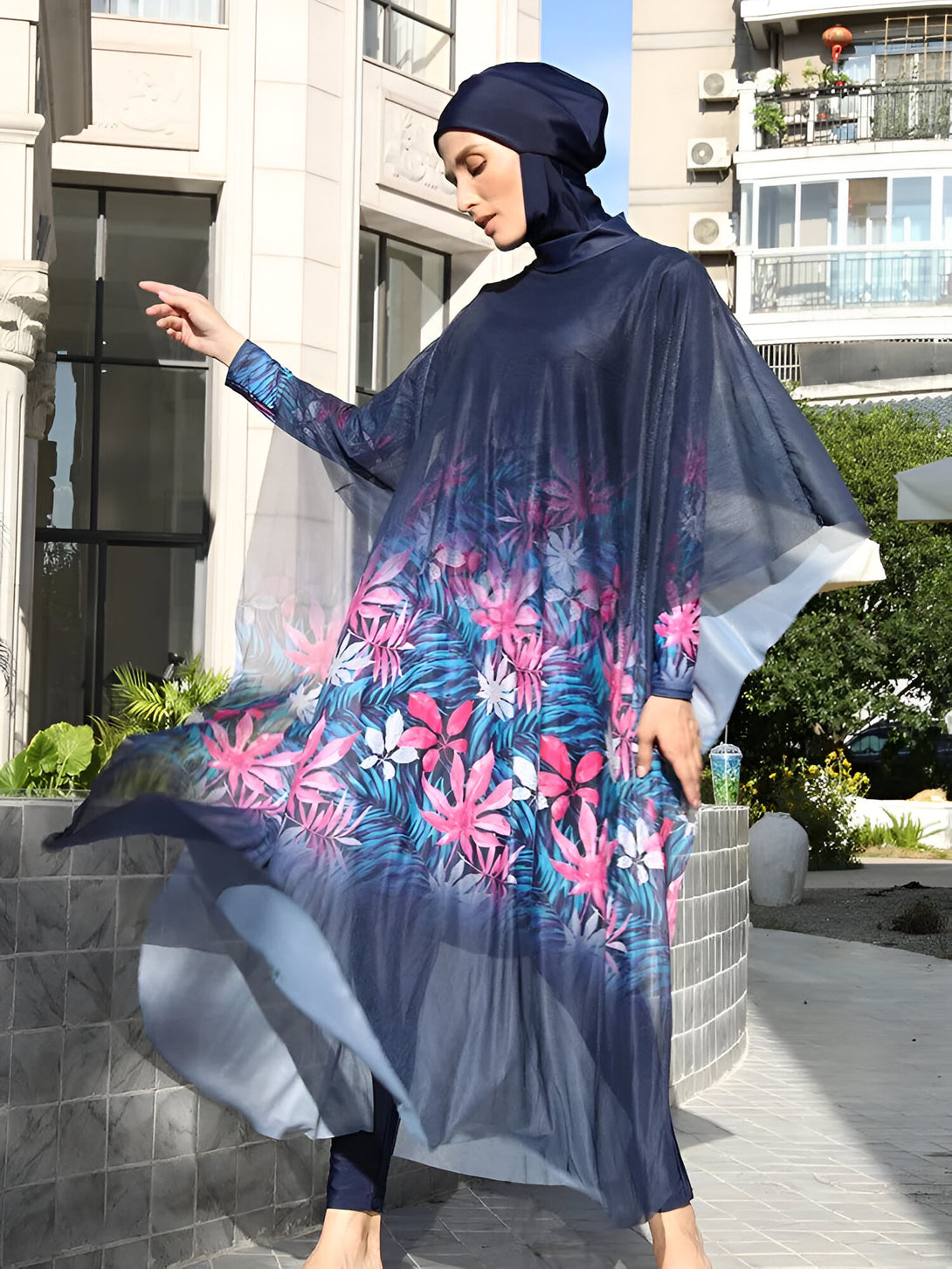 Une femme portant un maillot de bain musulman bleu marine à imprimé floral. Le maillot de bain comprend une robe longue et un hijab assorti. La femme est posée sur un fond urbain.