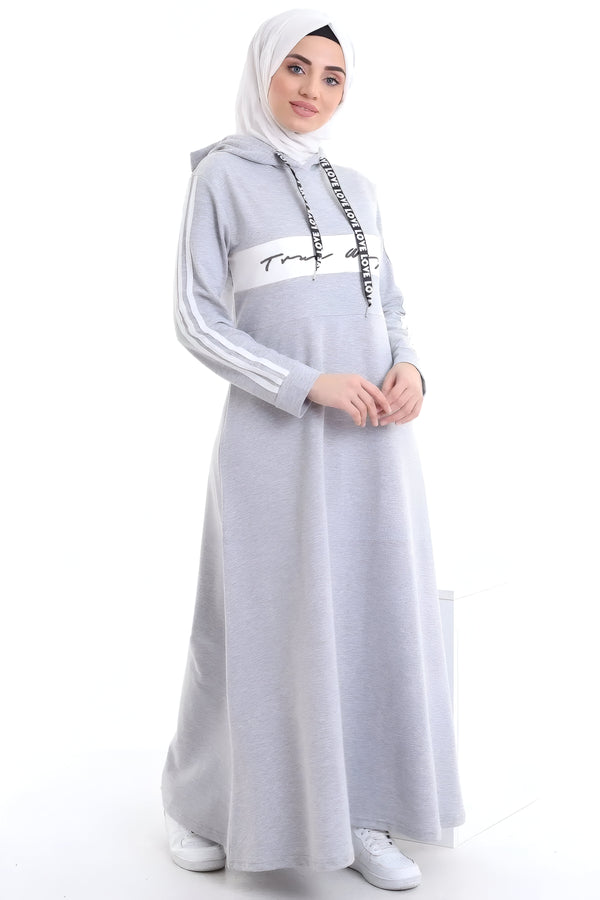 Une femme portant une robe grise à capuche avec des détails blancs et un hijab blanc. La robe a des manches longues et une coupe ample. Elle porte des baskets blanches.