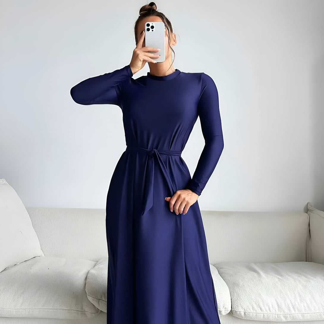 Une femme porte une robe longue bleue marine avec une ceinture à la taille, élégante et confortable, idéale pour les occasions spéciales ou au quotidien.