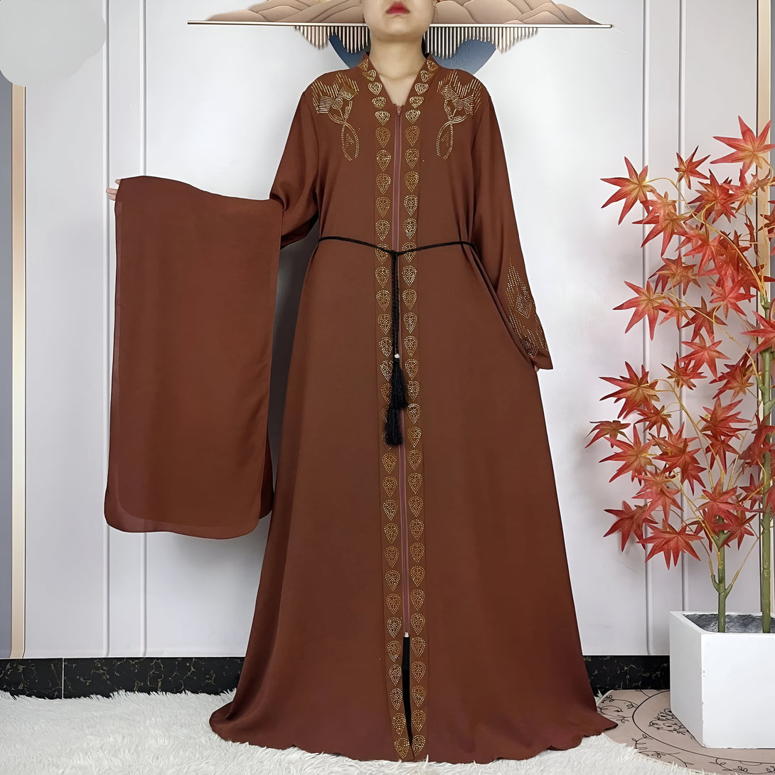 Une femme porte une abaya marron avec des détails dorés et une ceinture noire. L'abaya est décorée de pierres brillantes et a une fente sur le devant. Elle porte également un foulard marron.