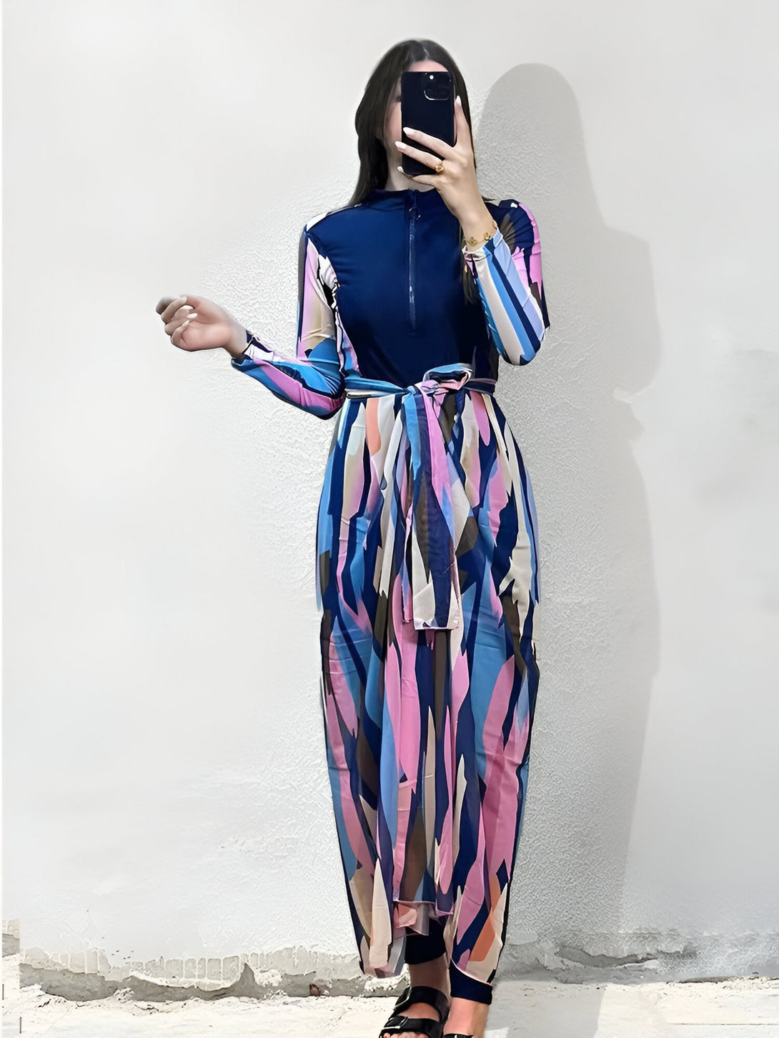 Une femme porte une robe de bain musulmane longue à manches longues, avec un imprimé géométrique bleu, rose et beige. La robe a une ceinture assortie. La robe est parfaite pour la natation ou la détente à la plage.