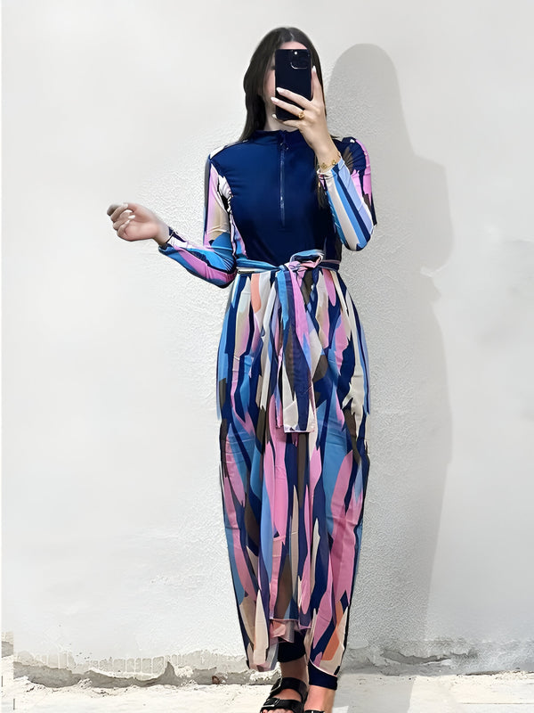 Une femme porte une robe de bain musulmane longue à manches longues, avec un imprimé géométrique bleu, rose et beige. La robe a une ceinture assortie. La robe est parfaite pour la natation ou la détente à la plage.