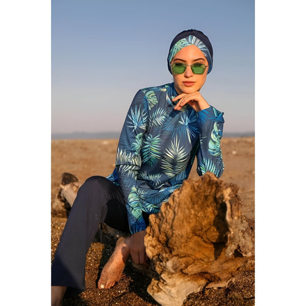 Une femme portant un hijab et un maillot de bain islamique à imprimé feuilles tropicales assis sur un tronc d'arbre à la plage.