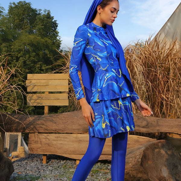 Une femme portant un hijab et un maillot de bain musulman bleu à imprimé marbré, debout devant un banc en bois dans un jardin.