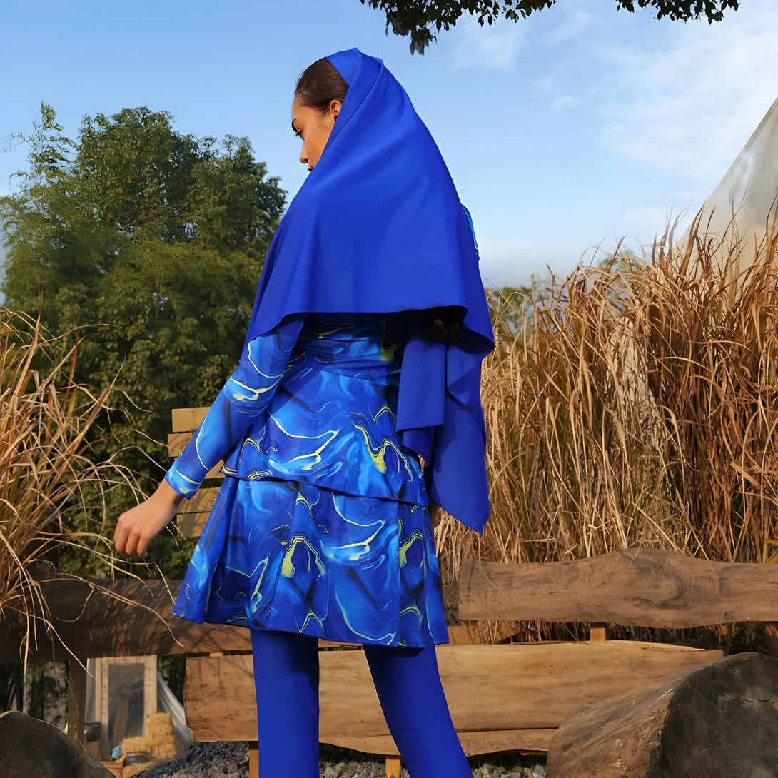 Une femme portant un hijab bleu et une tenue de bain islamique bleue à motif marbré, debout devant un banc en bois.