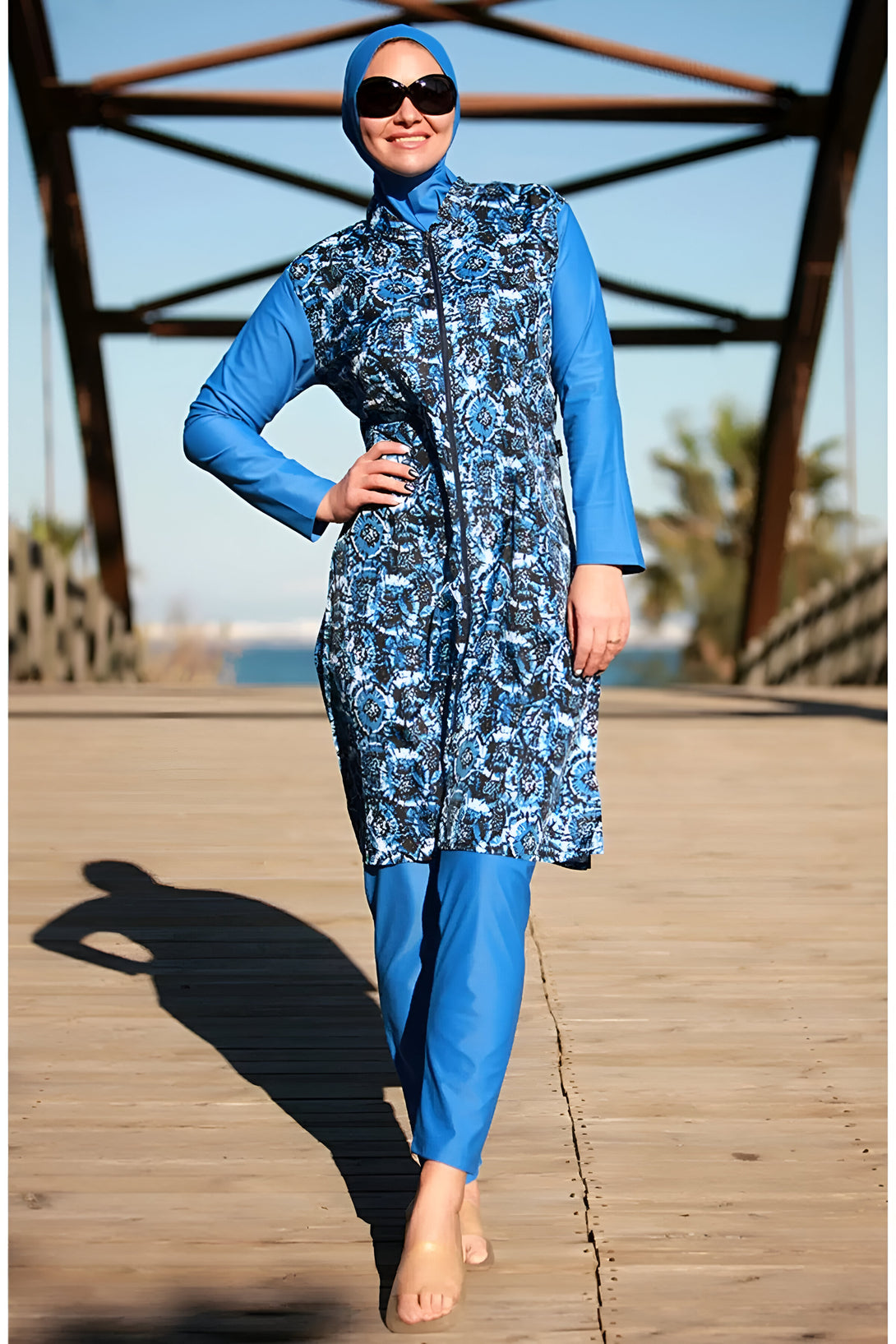Une femme portant un maillot de bain musulman bleu et blanc à motifs sur la plage. Elle porte un hijab et des lunettes de soleil.