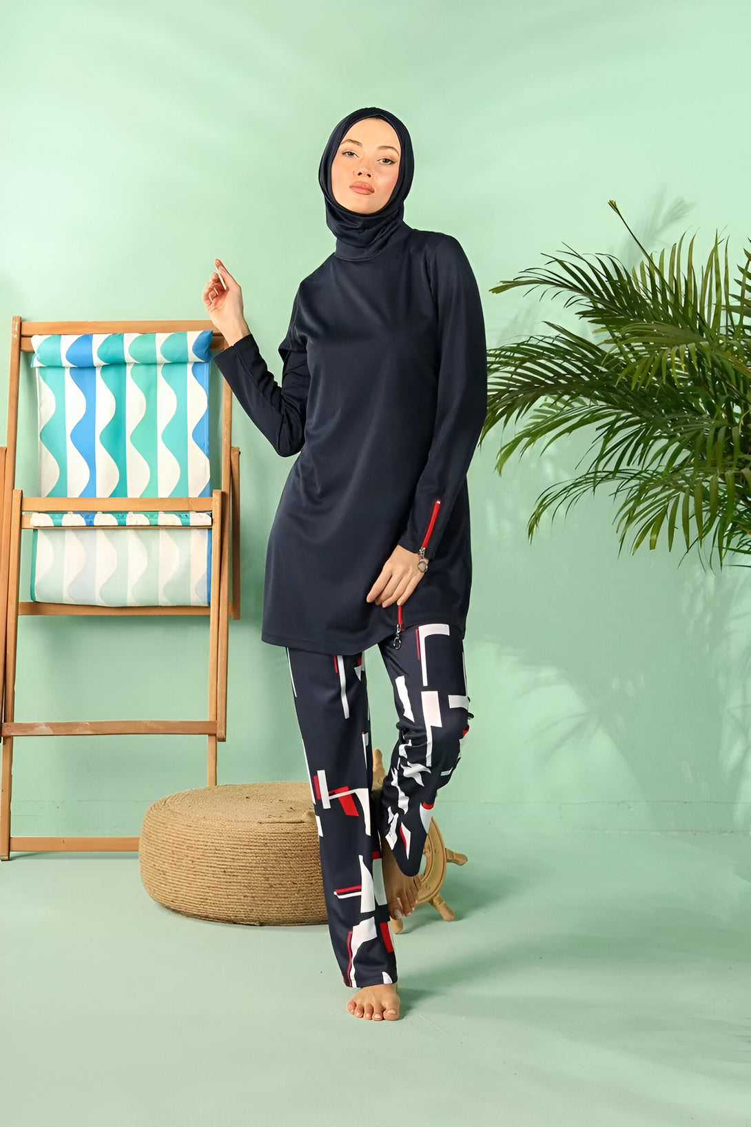 Une femme portant un maillot de bain musulman bleu marine avec des pantalons à motifs géométriques rouges et blancs. Elle se tient debout devant un fond vert avec un transat en bois et un pouf en jute.