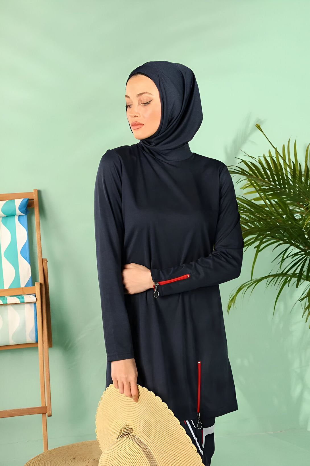 Une femme porte un maillot de bain musulman bleu marine avec une fermeture éclair rouge sur les manches. Elle porte un hijab assorti et tient un chapeau de paille.