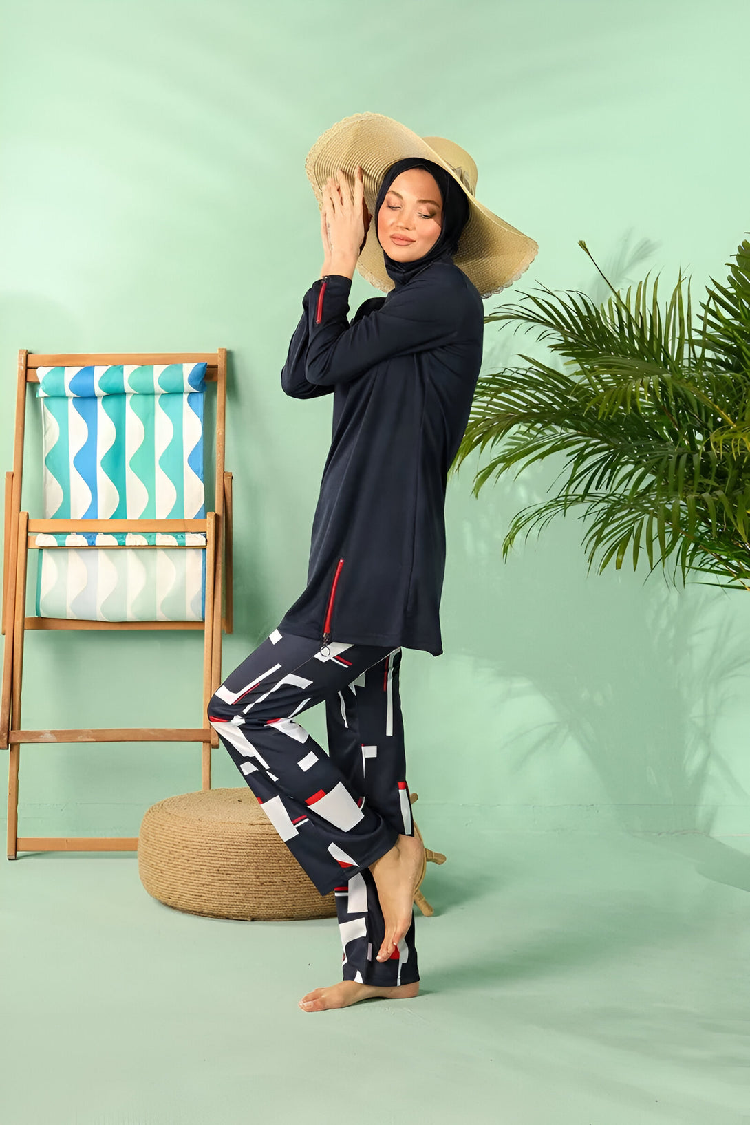 Une femme portant un maillot de bain musulman bleu foncé avec un pantalon imprimé géométrique noir et blanc. Elle porte un chapeau de paille et se tient à côté d'une chaise de plage et d'un palmier.